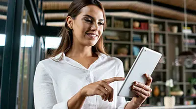 A woman in a white blouse smiling and pointing at a tablet in her hand. 